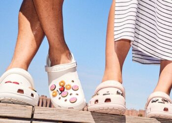 Zwei Personen tragen weiße Crocs mit buntem Motiv und gehen nebeneinander auf sonnigem Untergrund.