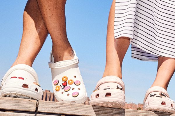 Zwei Personen tragen weiße Crocs mit buntem Motiv und gehen nebeneinander auf sonnigem Untergrund.