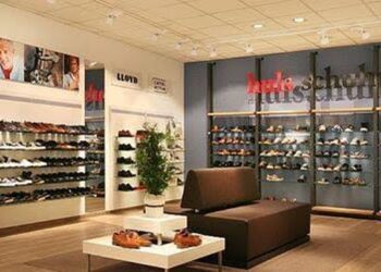 Moderne Schuhgeschäft-Innenansicht mit beleuchteten Schuhregalen an den Wänden, zentralem Holztisch und großflächigen dunklen Vitrinen