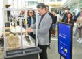 Mann in grauem Anzug betrachtet eine Ausstellungsvitrine mit Schuhmodellen auf einer Messe, Besucher im Hintergrund.