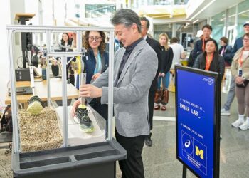 Mann in grauem Anzug betrachtet eine Ausstellungsvitrine mit Schuhmodellen auf einer Messe, Besucher im Hintergrund.