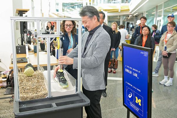 Mann in grauem Anzug betrachtet eine Ausstellungsvitrine mit Schuhmodellen auf einer Messe, Besucher im Hintergrund.