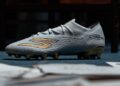 Weiß-goldener Fußballschuh mit strukturierter Oberfläche auf grauem Untergrund, Seitenansicht.