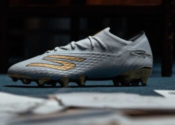 Weiß-goldener Fußballschuh mit strukturierter Oberfläche auf grauem Untergrund, Seitenansicht.