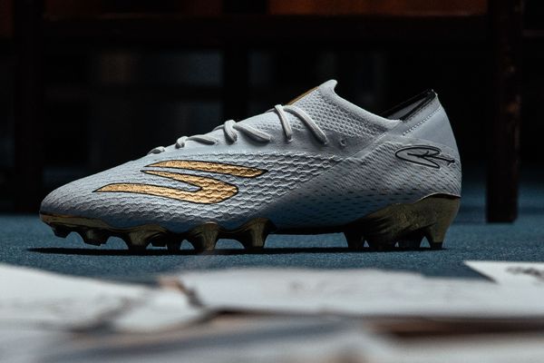 Weiß-goldener Fußballschuh mit strukturierter Oberfläche auf grauem Untergrund, Seitenansicht.